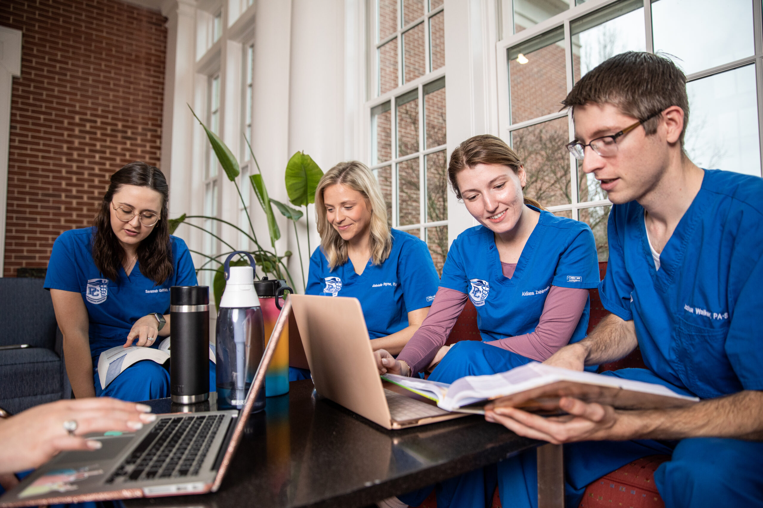 Presbyterian College PA students in blue scrubs studying together with laptops and textbooks in a bright campus space with large windows and natural light.