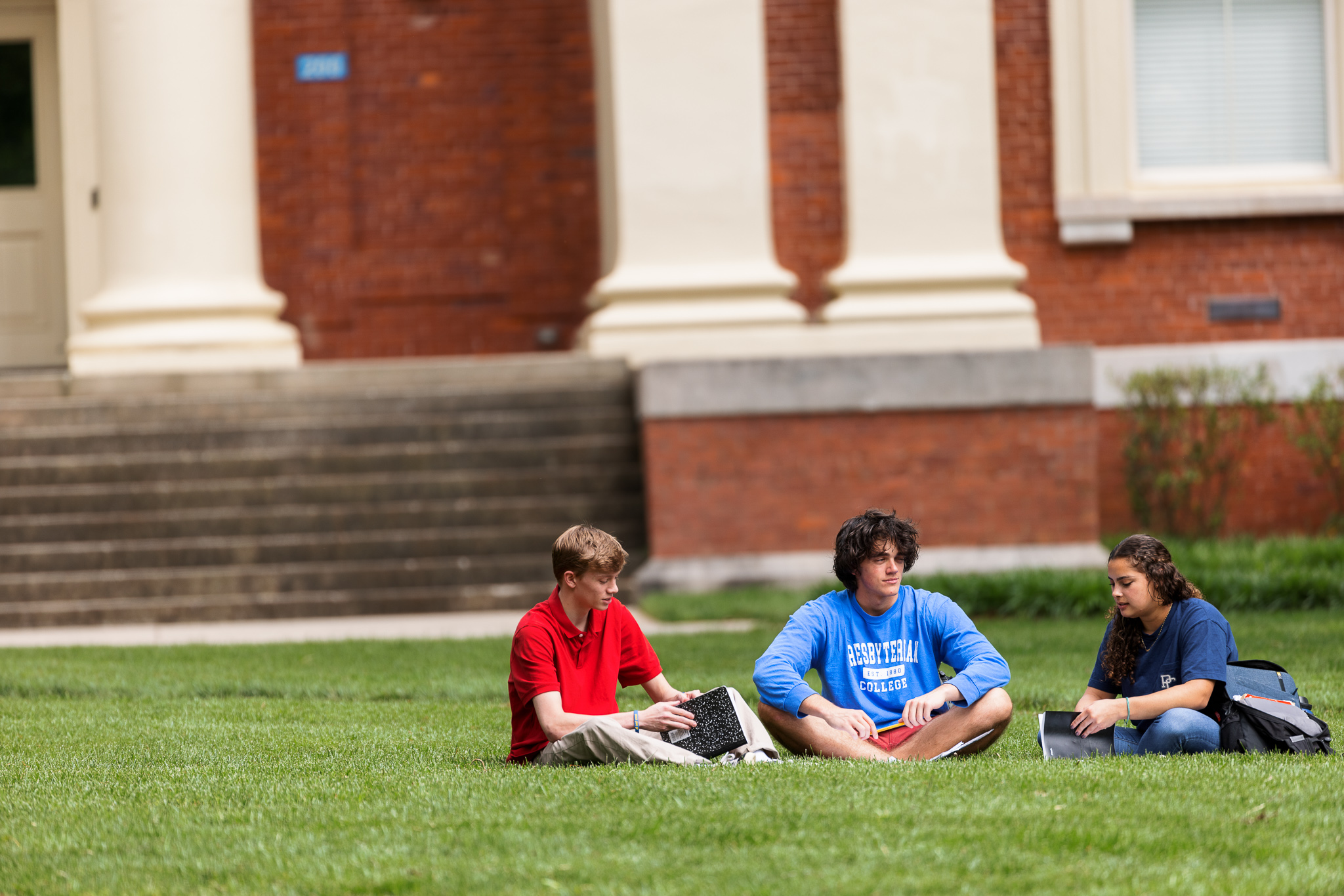 Three Presbyterian College students studying on the lawn in front of a historic brick campus building with white columns.